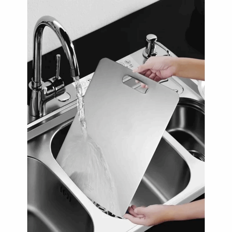 Person washing a stainless steel cutting board under running water in a kitchen sink.
